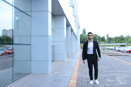Young handsome businessman Arab Muslims is in the business center in black elegant dress on building background. Businessman man male looking at time walking goes and smiling.の写真素材