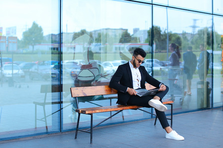 Entrepreneur uses laptop wearing sunglasses. Young handsome businessman arab muslims in business center wearing dressed in black elegant suit on building background.の写真素材