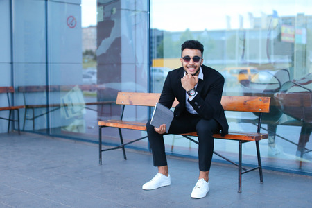 Entrepreneur smiling man  puts on sunglasses uses tablet and looks at. Young handsome businessman arab muslims in business center wearing dressed in black elegant suit on building background.の写真素材