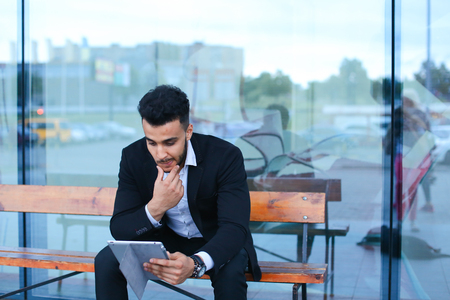 Entrepreneur smiling man  puts on sunglasses uses tablet and looks at. Young handsome businessman arab muslims in business center wearing dressed in black elegant suit on building background.の写真素材