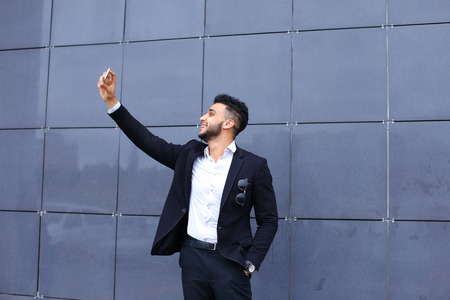 Entrepreneur takes makes selfie man male looking at camera and smiling talks on mobile smart phone. Young handsome businessman   in business center wearing dressed in black elegant suit on building background.の写真素材