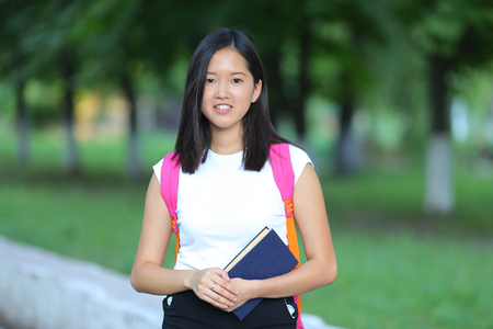 Girl female teenager with black hair in a bob white east Asian woman green park with a backpack background. Lady holds, dials, push your finger, smiling, looking into the camera, talking on the phone happy portrait.の写真素材