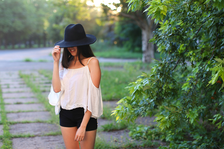 female nature keeps his hat summer sun sunset looks pleased. azitka woman black hair white shirt black shorts dancing around the trees looking at the camera. portrait. spinningの写真素材