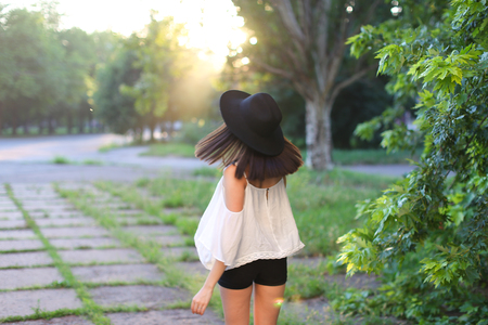 female nature keeps his hat summer sun sunset looks pleased. azitka woman black hair white shirt black shorts dancing around the trees looking at the camera. portrait. spinningの写真素材