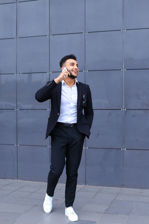 Entrepreneur man male looking at camera and smiling talks on mobile smart phone. Young handsome businessman  in business center wearing dressed in black elegant suit on building background.の写真素材