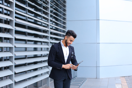 Young handsome businessman   is in the business center in black elegant dress on building background. Businessman man male looking waiting concept and smiling.の写真素材