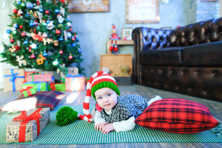 Cute baby lying on plaid among gifts on background of Christmas tree in funny hat with bubo. Concept of baby, assistant Santa, Christmas mood, giving presents, gifts, Christmasの写真素材