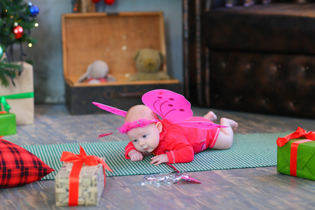 Little baby lying on plaid in fairy costume with pink wings and rim on head against background of New Year's decor in studio among the gifts. Concept of baby, assistant Santa, xmas, Christmas mood, giving presents, gifts, happy Christmas.の写真素材