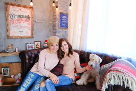 Daughter with mother sitting together on brown leather couch next to dog, small terrier fluffy, smile, mother hugs daughter,girl petting dog on background of large window with curtains and gray walls with posters in New Year's studio. Woman dressed in palの写真素材