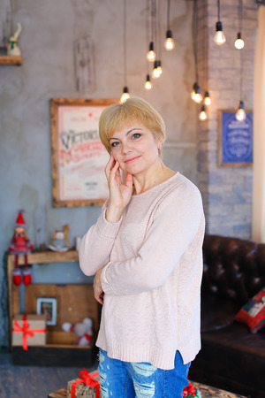 Lovely lovely female woman smiles little smile, standing and posing right in front of camera on background of gray walls, decorations and glowing lights in festive Christmas photo studio. Woman dressed in a pale pink knitted sweater and blue ripped jeans.の写真素材
