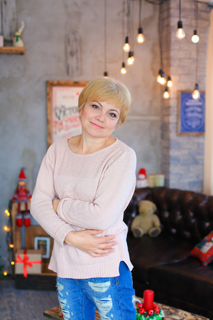Lovely lovely female woman smiles little smile, standing and posing right in front of camera on background of gray walls, decorations and glowing lights in festive Christmas photo studio. Woman dressed in a pale pink knitted sweater and blue ripped jeans.の写真素材