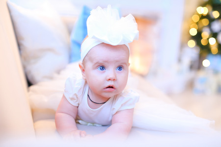 Little infant girl lying on sofa in white dress and white rim with flower on head on background luminous Christmas tree and white stone fireplace. Concept of little Princess, Christmas holidays, New Year's Eve, family celebration.の写真素材