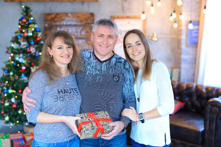 Lovely family smiling, posing at camera, holding gifts in hands and hugging each other for family photo in studio on New Year background luminous Christmas tree. Woman dressed in gray sweater and blue jeans, man dressed in dark blue sweater and blue jeansの写真素材