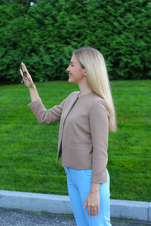 Charming Lady European appearance rejoices acquisition machine or your own apartment, overflowing emotions girl smiling broadly and showing keys. Women with long blonde hair dressed in bright blouse, beige jacket and blue pants, standing on background of の写真素材