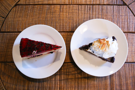 Top view of piece of cherry cheesecake with jelly and chocolate cake decorated with whipped cream on white round plates, which stand on round wooden table in cafe. Concept of delicious food, pleasure, confectionery, restaurant business.の写真素材