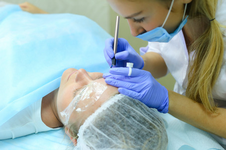 Young blonde woman professional, engaged in reconstruction work responsibly, eyebrows manual method performs procedure, using thinnest needle and maniples, draws most natural eyebrow, side by side on couch and ready for client procedure in cosmetic white の写真素材