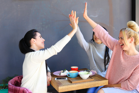 Three young attractive girls girlfriends rejoice, high five to each other, chatter, gossiping, share secrets, laughing, smiling and sitting on chair at table on background of wall and trees in coffee shop. Girls with black hair dressed in  white and gray の写真素材