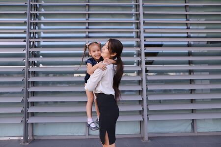 Charming modern female woman, young mother, successful business woman squatted down next to daughter, smile at each other, hug with girl, small child hugging her mothers neck and kisses on cheek, posing for camera, girl holding baby girl on background of の写真素材