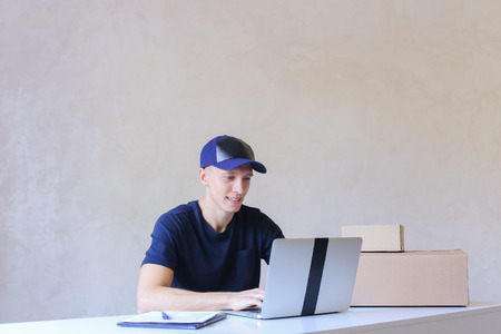 Young Postman Sits at Table Typing at Laptop. Courier Company Accepts Orders Every Day Different Types and Sizes Young Deliveryman European Appearance in Black T-Shirt and Cap Sitting at White Table Typing on Computer and Takes Pen Writing Down Informatioの写真素材