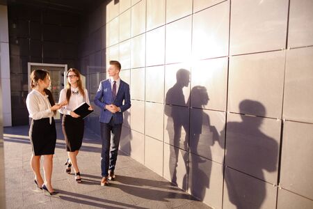 Two elegant successful girls and handsome male man young office workers, businessmen, students talk with each other, laughing and smiling, discussing working moments, hand-held instruments and stand against background of business center outdoors. Girls wiの写真素材