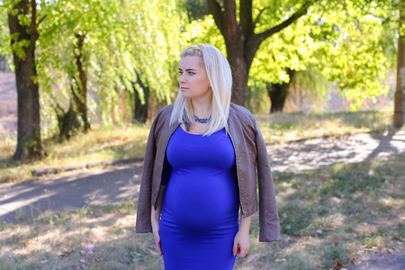 Charming Lady Being Prepared to Become Mother. Young Woman Photographed For Album of Family Photographs or Advertising Clothes For Pregnant Women, Waiting For Her Husband in Park to go Along to the Restaurant For Dinner. Blonde With Hair Braided in Pigtaiの写真素材