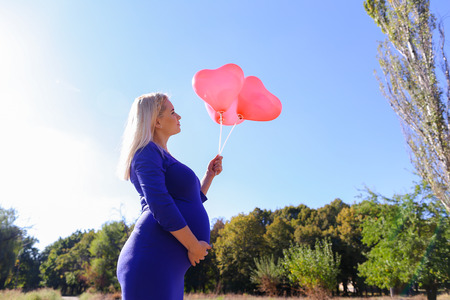 Lovely Young Adult Dirl in Family Way. Blond Woman Luxuriates on Sun and Holds in Hand Three Pink Balloons in Form of Heart, Looks at It, With Care and Affection Stroking Tummy and Enjoys Clear Sunny Day and Life. Lady in Large Park With Trees in Outdoorsの写真素材