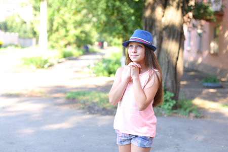 Pretty cute female girl child smiles, make wish, dreamily looking away, holding hands near face and standing in yard on street. Girl dressed in pink shirt, printed shorts, dark blue hat with brim. Concept of childhood, desire, dream, summer holidays, goodの写真素材