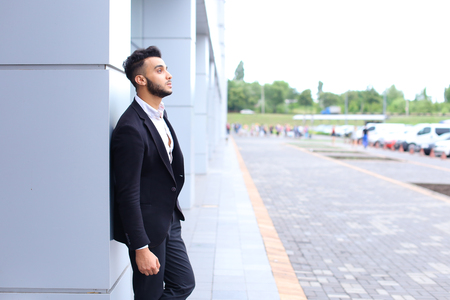 Young handsome businessman Arab Muslims is in the business center in black elegant dress on building background. Businessman man male looking at time walking goes and smiling.の写真素材