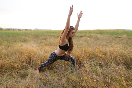 Young female fitness instructor performs warm-up exercise, stretching and holding focuses on correct execution poses, photographed for advertising of catalog womens sportswear or sports complex in wide field outdoors. Sportswoman with curly brown hair dreの写真素材