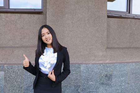 Pretty diligent student female asian girl  looks straight at camera, holds thumbs up, shows gesture indicating cool, smiling against backdrop of business building. Woman with black hair, dressed in white blouse,classic jacket and skirt.Concept of student の写真素材
