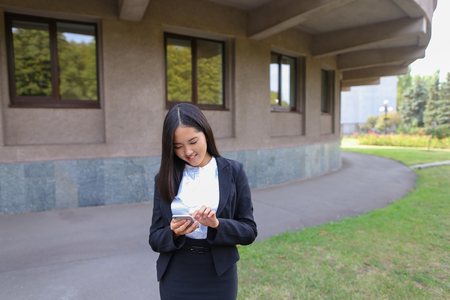 Beautiful cheerful young female international student girl holding phone, smiling, laughs and chatters. Woman with black hair, dressed in white blouse, classic jacket and black skirt. Concept of advertisement of mobile phone, technology, student everyday の写真素材