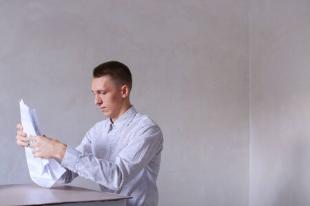 Entrepreneur adds documentation and completes work day. Men with short hair and blond hair sitting on chair at white table in office and dressed in white classic shirt. Concept of businessman office work, sedentary routine work documentation, successful yの写真素材
