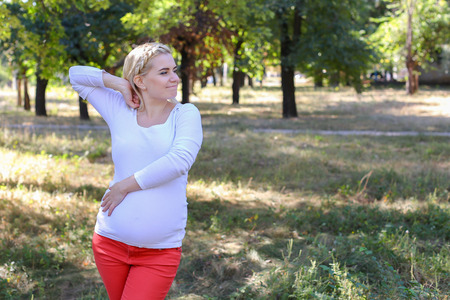 Lovely Woman and Future Mother Prepares For Birth of Baby, Tired and Trying to Relax, Posing For Photos on Memory and Stands in Green Sunny Park Outdoors in Daytime. Girl With Blond Hair, Braided in Braid in Front Dressed in White Blouse With Long Sleevesの写真素材