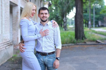 Young couple standing next to each other, smiling, posing and hugging on street. Girl turned sideways in profile and dressed in midi gray dress, guy standing straight and looking directly into camera, he dressed in blue jeans and white striped shirt. Concの写真素材