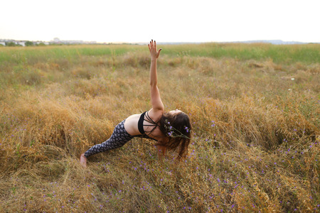 Young female fitness instructor performs warm-up exercise, stretching and holding focuses on correct execution poses, photographed for advertising of catalog women's sportswear or sports complex in wide field outdoors. Sportswoman with curly brown hair drの写真素材