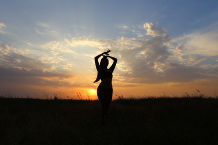 Female with beautiful figure takes yoga postures, gymnastics and ballet spends stretching and meditating, posing for advertising shoot fitness studio, healthy energy drink, dancing belly dancing among wide field on background of yellow sunset and beautifuの写真素材