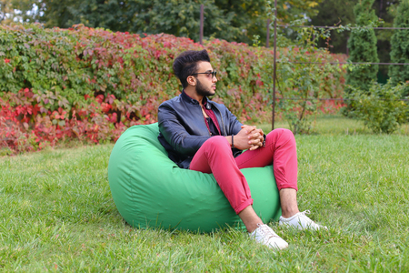 Arabian Young Man Sitting and Resting From City Bustle in Soft Green Chair, smiles and Looks Around. Handsome Guy Has Dark Hair and Black Beard and dressed in Maroon Shirt Over Black Leather Jacket and Bright Pink Jeans and White Sneakers Accessories Fromの写真素材
