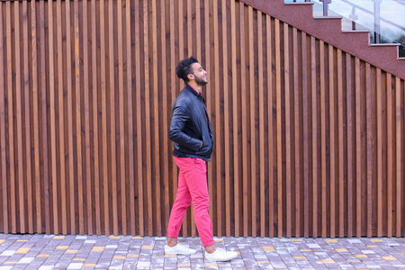 Handsome Young Adult Muslim Man, Businessman Walks Slowly Along Stairs, Looks Around and Smiles, Holds Hands in Pockets of Jackets, Advertises Clothing on Background of Wooden Stairs Near Restaurant Outdoors. Confident Arab Guy With Dark Hair Dressed in Bの写真素材
