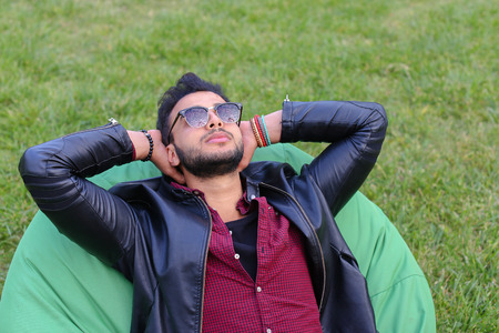 Portrait of Young Arab Masculine Man Student, Which Lies on Soft Green Chair, Dreams, Meditate, Thinking About Something, Smiling and Looking at Heaven, Hands Behind Head on Background of Green Grass Outdoors. Stylish Guy With Dark Hair Dressed in Black Tの写真素材