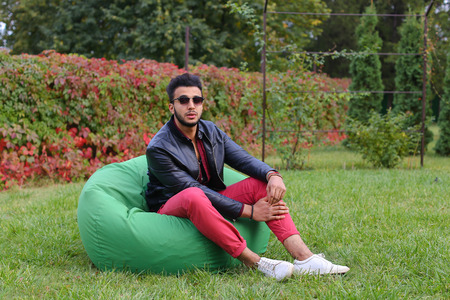 Arabian Young Man Sitting and Resting From City Bustle in Soft Green Chair, smiles and Looks Around. Handsome Guy Has Dark Hair and Black Beard and dressed in Maroon Shirt Over Black Leather Jacket and Bright Pink Jeans and White Sneakers Accessories Fromの写真素材