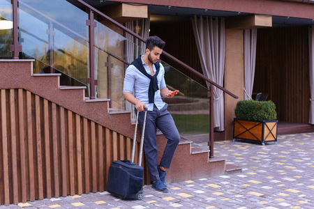 Young man businessman Arabian beautiful appearance is black travel bag on small wheels and is waiting for taxi, on journey or business trip, using phone and check their seats in airplane online, reserve hotel, one leg held up other, and based on stairs neの写真素材