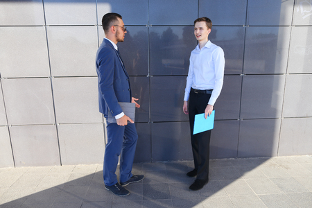 Two beautiful active young male businessman guys discussing important issues, make decisions, solve problems, share business ideas, smiling, holding in hands colored folders and stand opposite each other near gray wall of modern business center on outdoorの写真素材