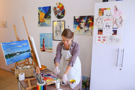 Artist attractive woman of European appearance writes oil painting, enjoying things what love , paint picture for sales and order, listening to music on headphones through phone, stands and works in bright art studio against background of wall hung with pの写真素材