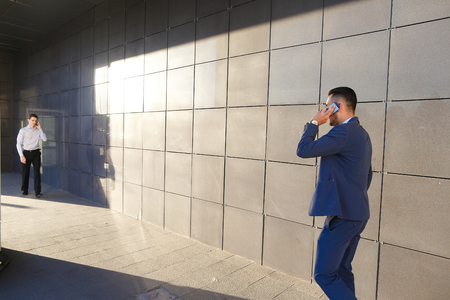 Young perspective male men, students,go to meet each other, greet one another, shake hands, talking on phone, stop and talk, walk fast step, hurry, running late on background of wall of business center outdoors .One of guys dressed in blue classic suit, wの写真素材