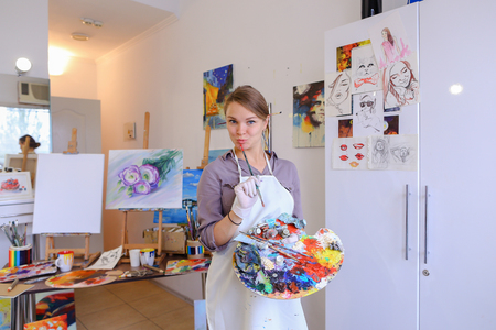 Cute female painter posing, laughing and fooling around in front of camera and smiling with brushes and colorful palette in hand, standing in studio with white art paintings on walls. Girl European appearance advertises art school for adults and children,の写真素材