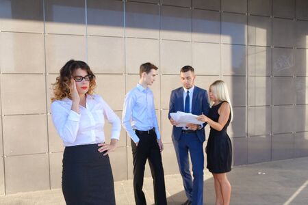 Pretty young perspective business woman, student holds on head from fatigue, rests, took break from work, holds paper and waving them, watching and stands on background of colleagues who discuss workflows, consider paper near of business center on street.の写真素材