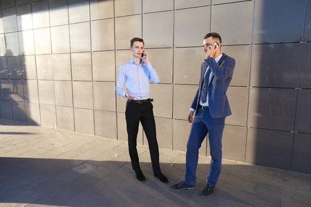 Two goodly active confident young male men, businessman, students talking on phone, solve important questions, make plans, arranging meetings and stand on background wall of modern business center outdoors. One of guys dressed in blue classic suit, white の写真素材