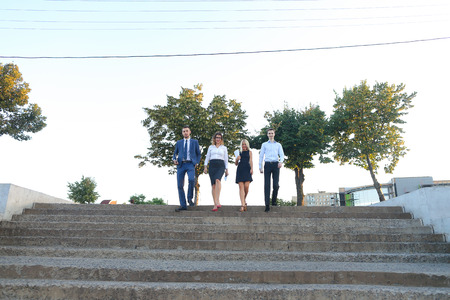 Young modern confident people, two boys and two girls, perspective entrepreneurs, students smiling, hold in hands gadgets, tablet, smartphone and papers, documents, go down stairs outdoors. One of guys dressed in blue classic suit, white shirt with tie, sの写真素材