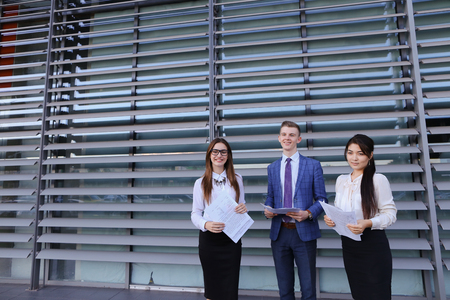 Three energetic young beautiful novice businessman, educated students, office workers, two cute girls and man, handsome guy posing and smiling for photograph in camera, keep documentation in hands, folders with working materials, taken to promotional videの写真素材