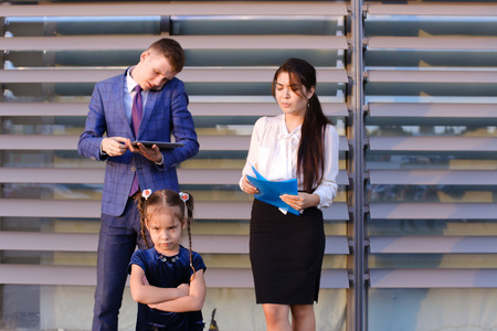 In  foreground stands and frowns, angry with arms crossed in front of little girl, child,  in background successful young handsome mans man, young father and modern woman, young mother hold gadgets in hand, mobile phones and talking to them, solve operatiの写真素材